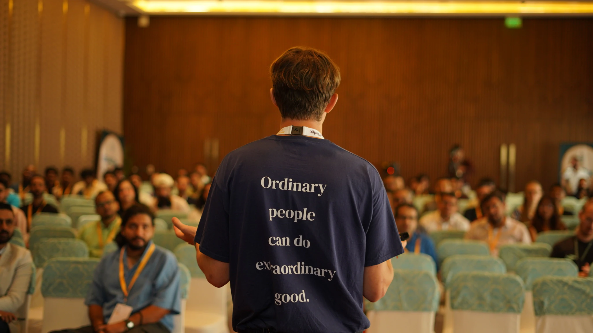 A Giving What We Can member giving a talk, wearing a T-shirt that reads “Ordinary people can do extraordinary good.”